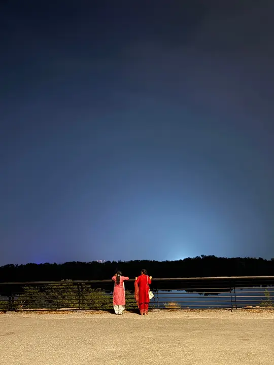 Two people in red and pink kurtas at a railing by the water under a deep blue night sky